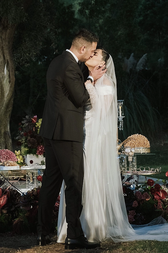 Wedding kiss as the bride and groom kissing in a close embrace, veil flowing, by a glass decor table with florals on a garden lawn