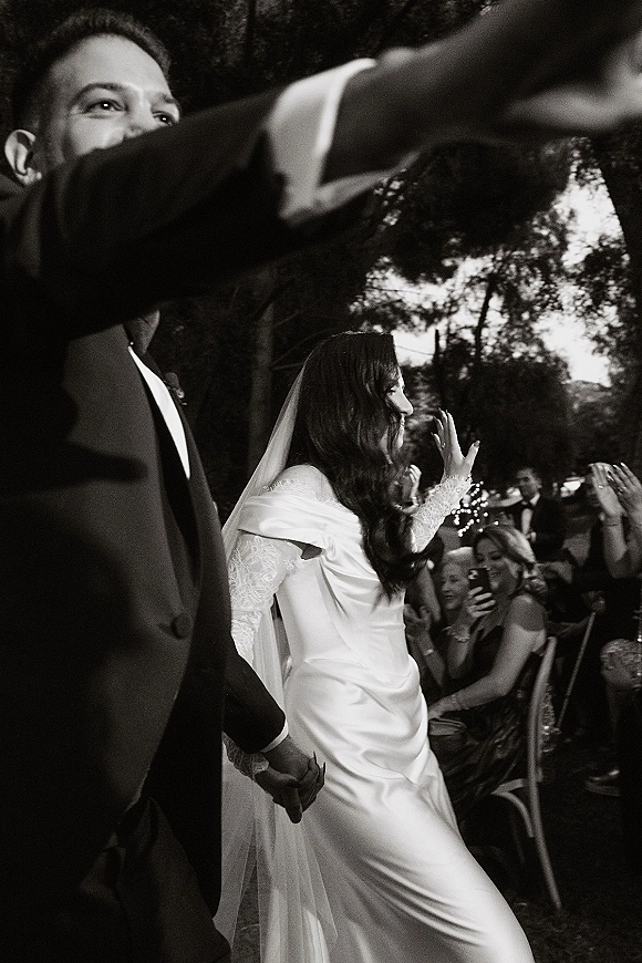 Wedding recessional as bride and groom walk hand in hand up the aisle, her lace sleeves and veil in view, guests cheering under string lights
