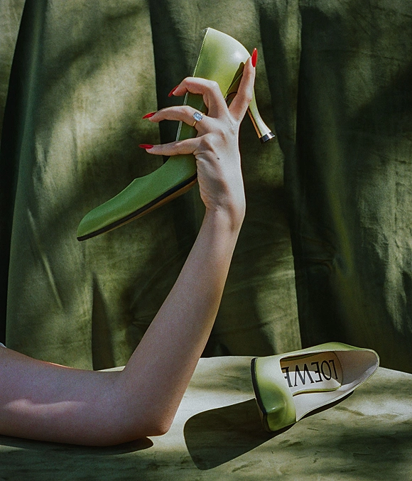 Bridal shoes in green wedding shoes style on draped green fabric, with engagement ring, red manicure, and gold heel detail in shadows