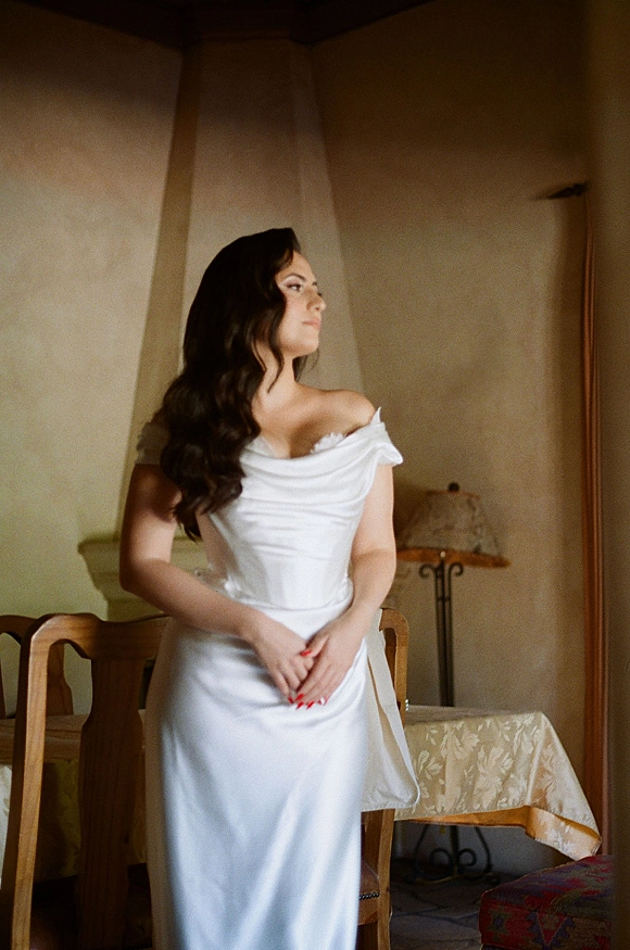 Bridal portrait of a bride looking away in an off the shoulder satin gown, hands clasped, long wavy hair in a warm indoor room with chairs and a lamp
