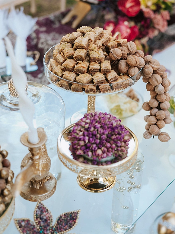 Wedding dessert table with baklava dessert display on gold cake stand, walnuts, white taper candle, glass cloche, and florals on glass tabletop