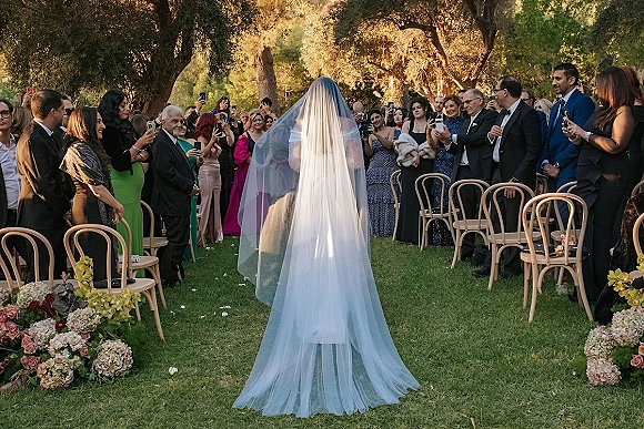 Wedding processional as the bride walking down aisle with a long cathedral veil, rose petals and flowers on a sunlit garden lawn