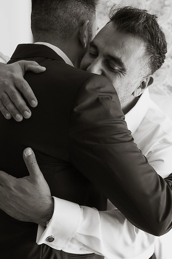 Groomsmen hug tightly in a candid groom and best man moment, suit jackets and cufflinks visible against a simple light wall backdrop