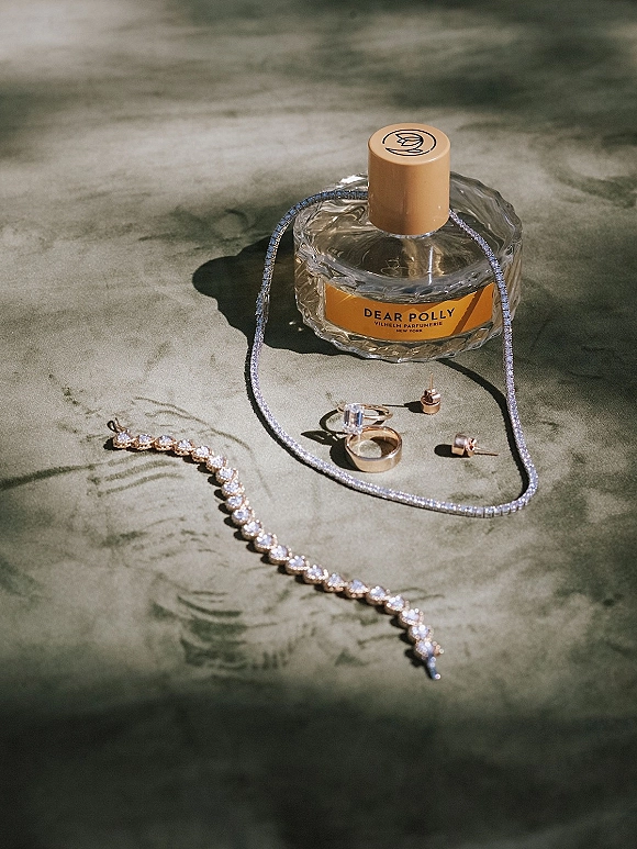 Wedding jewelry flatlay featuring an engagement ring, gold wedding band, stud earrings, tennis bracelet and necklace beside perfume on a sunlit stone surface