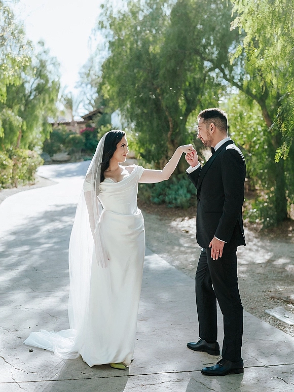 Couple portrait of bride and groom outdoors as he kisses her hand, her long veil and satin dress glowing on a sunlit garden walkway