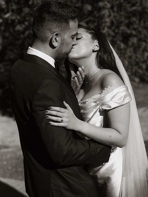 Wedding kiss in a black and white wedding photo, bride in off-shoulder gown and veil embracing groom in tuxedo amid blurred greenery