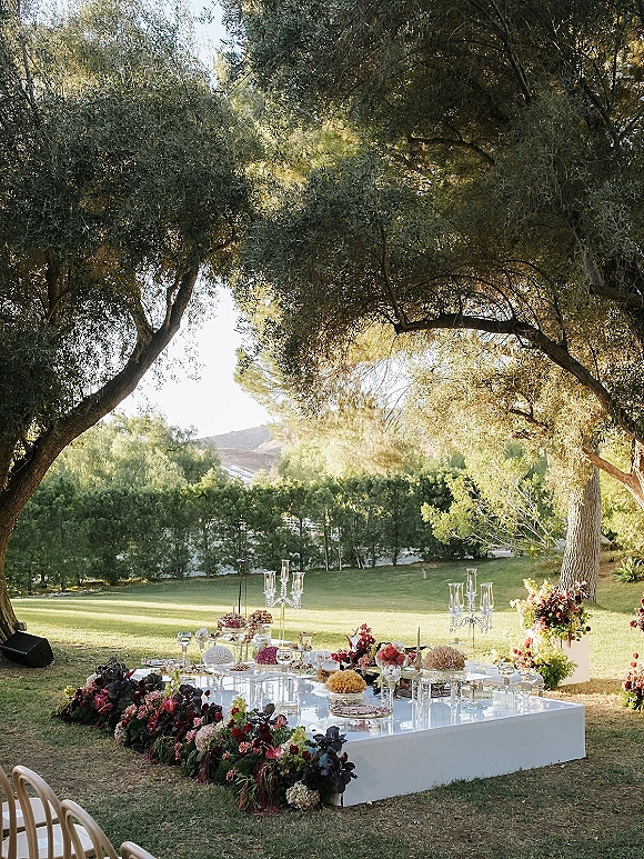 Ceremony altar decor on a white platform wedding altar with clear candelabras, taper candles, and lush florals on a sunlit lawn under trees