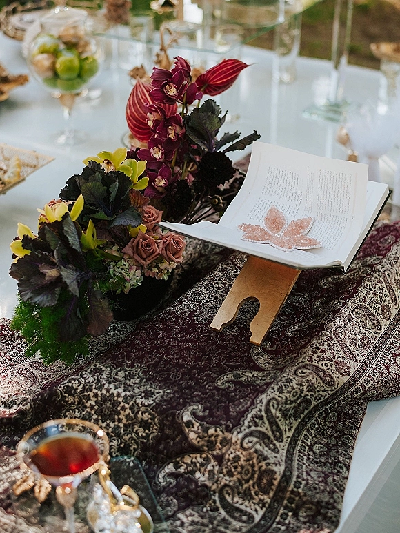 Wedding guest book on a guest book table, open on a wooden stand with orchid and rose florals, butterfly accent, and goblets outdoors