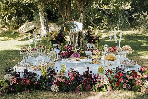 Reception dessert table with glass pedestal stands, crystal candelabras, and roses beneath an ornate gold mirror on a garden lawn
