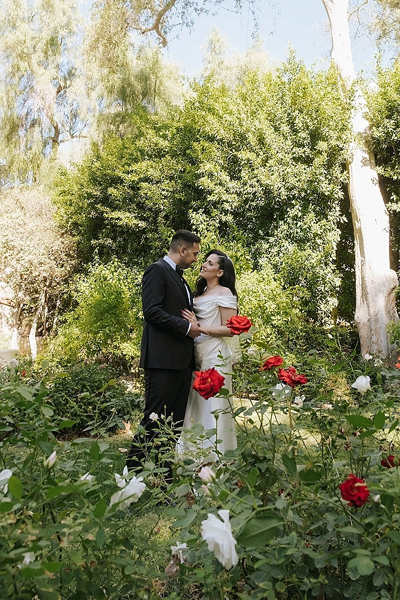 Couple portrait of bride in an off-the-shoulder gown and groom in black tuxedo holding hands beside red and white rose bushes in sunlight