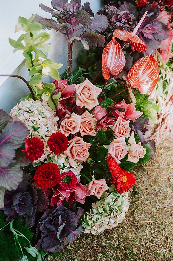 Wedding florals arranged as a wedding floral arrangement on grass by white drape, featuring roses, orchids, anthurium, hydrangea, and purple kale