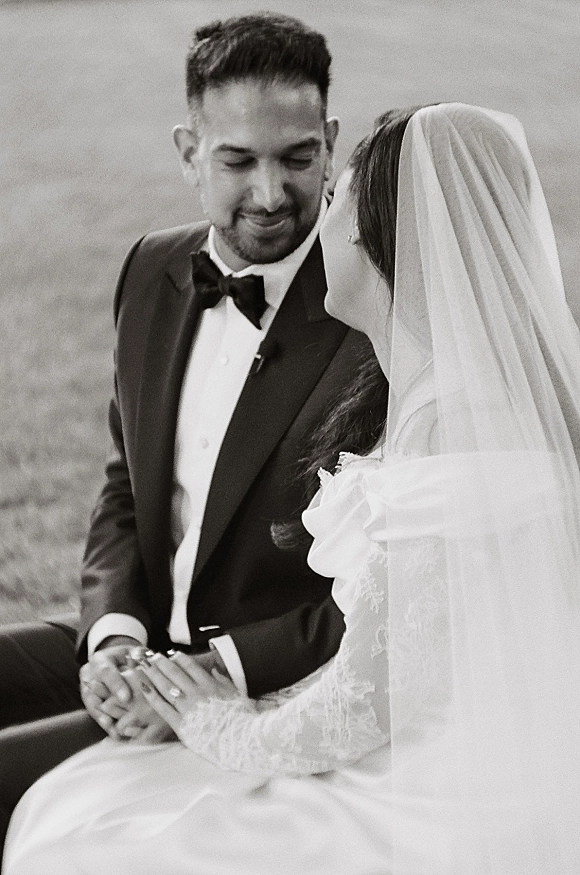 Couple portrait in a black and white wedding portrait, bride in veil and lace sleeves holding groom’s hand on a grass field