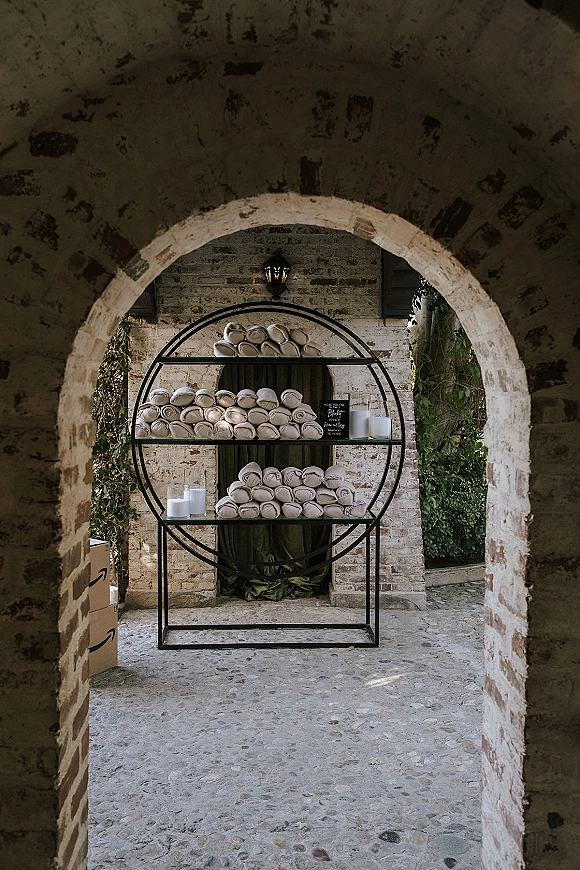 Wedding welcome towels and rolled towel display on a circular metal shelf with chalkboard sign and pillar candles by a brick archway