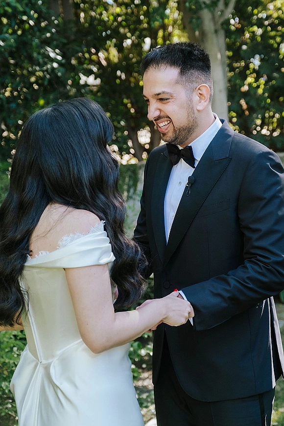 First look moment as bride in an off-the-shoulder lace-trim dress holds hands with groom in tuxedo, smiling in a sunlit garden
