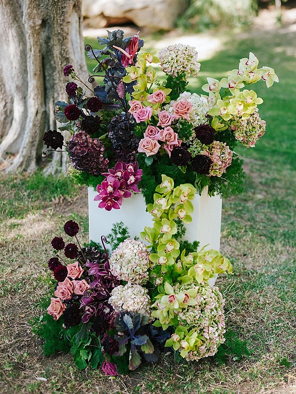 Wedding floral arrangement on a white pedestal plinth with ceremony pedestal flowers, orchids, roses, and greenery beside a garden tree trunk