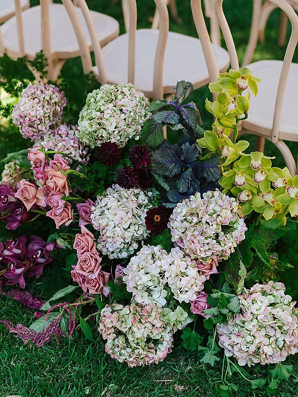 Wedding ceremony florals and aisle floral arrangement with hydrangeas, roses, orchids, and greenery beside white chairs on a grass lawn