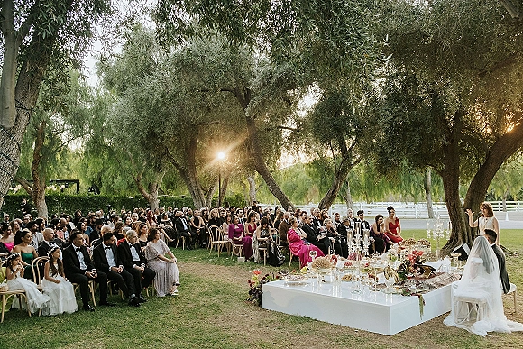 Wedding ceremony outdoors with guests seated under large trees, a white platform stage and candelabras glowing in warm sunset light