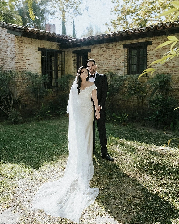 Couple portrait of bride and groom holding hands, her cathedral veil and long train on grass, posed by a brick house garden