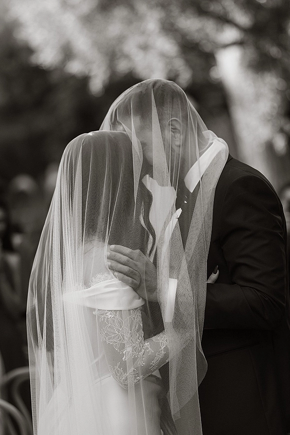 Wedding kiss as bride and groom embrace under a flowing veil, lace sleeves and tuxedo visible, with blurred trees behind
