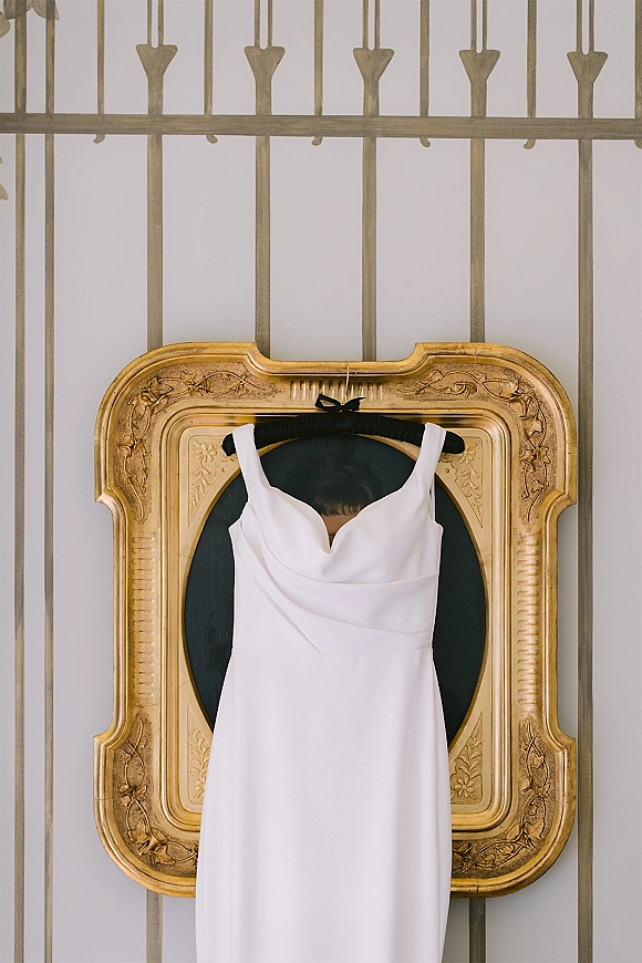 Wedding dress on a black hanger, simple wedding dress framed in ornate gold against a striped wall with metal railing