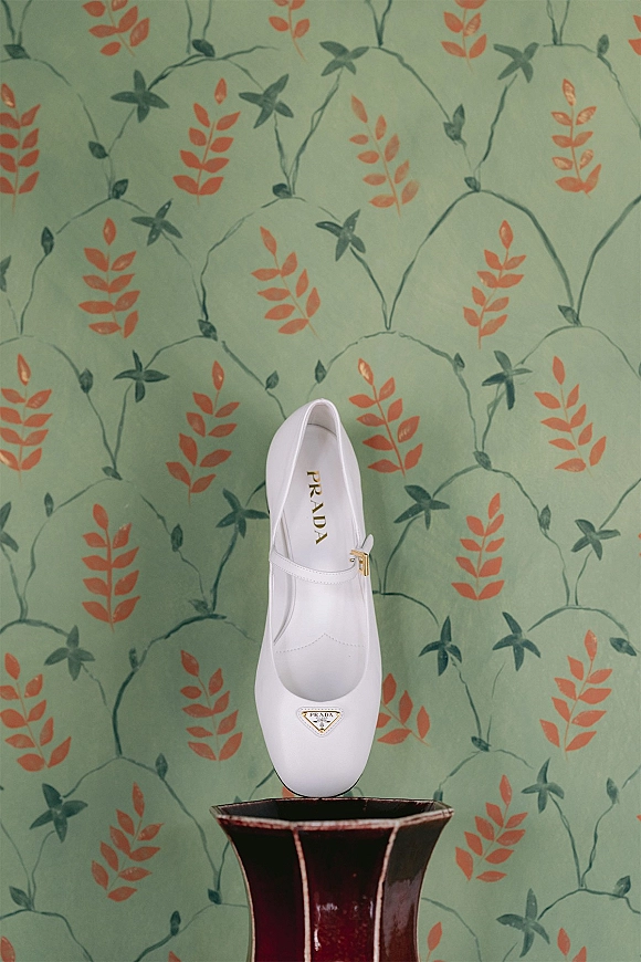Bridal shoes in white leather mary jane heels with a gold buckle strap and logo plaque against green floral wallpaper beside a vase
