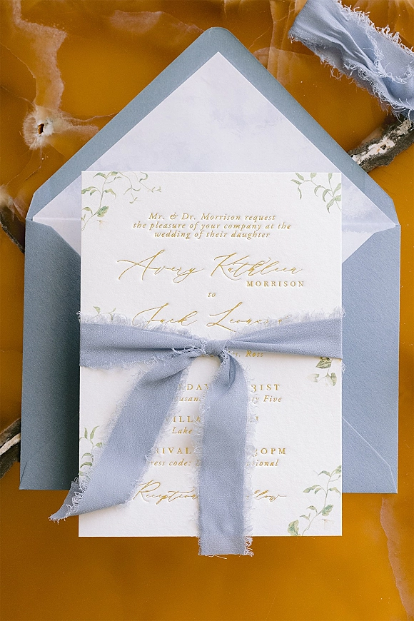 Wedding invitation suite with gold foil lettering and botanical illustration, styled with a blue envelope and ribbon on amber marble stone surface
