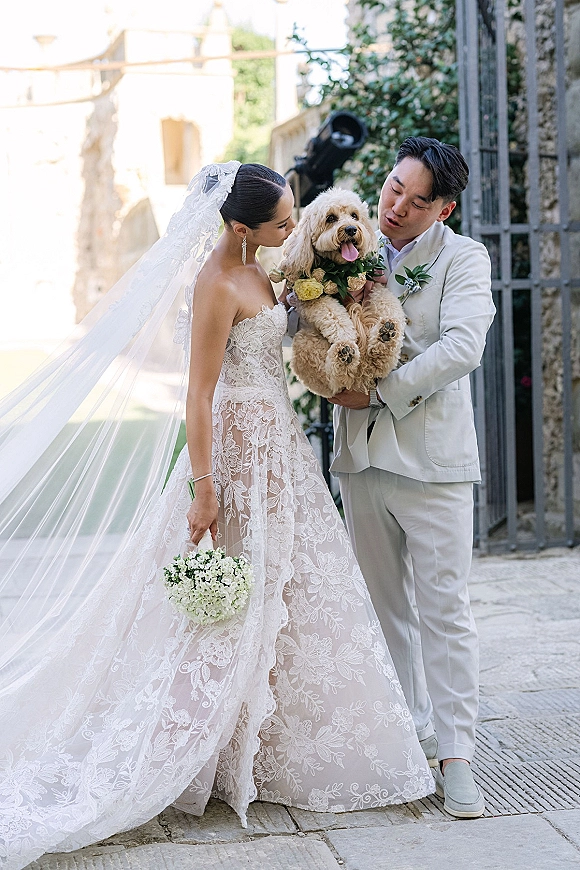 Couple portrait of bride and groom with dog, groom holding pup in floral collar as bride smiles in a stone courtyard gate backdrop
