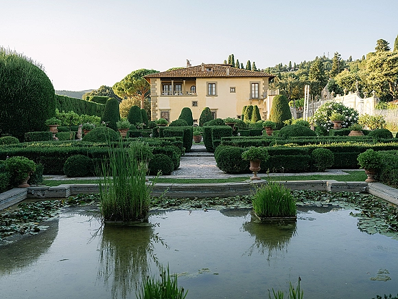 Garden wedding venue with manicured hedges surrounding a water lily pond, stone paths, and a villa backdrop under open sky