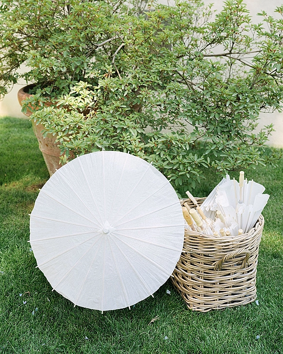 Wedding parasols in a wicker basket with white paper parasols arranged for guests on a green lawn beside a stucco wall