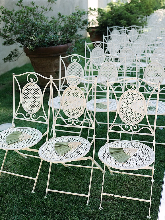 Ceremony seating with white wrought iron chairs and palm leaf fans arranged on a grass lawn beside potted greenery and a light wall