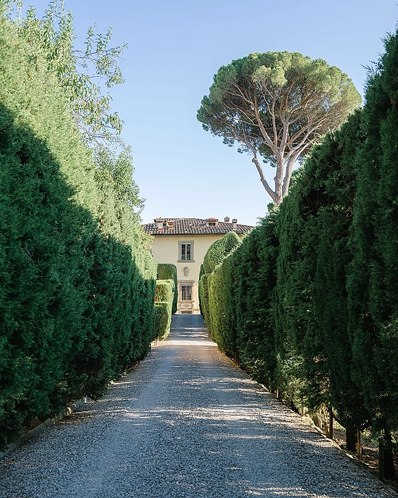 Wedding venue driveway with a gravel path and manicured hedges, tree lined driveway leading toward a villa under a clear blue sky