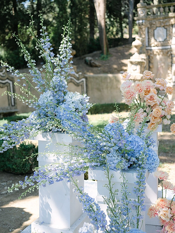 Wedding floral arrangement with ceremony pedestal flowers featuring blue delphinium and peach roses on white plinths in a garden courtyard setting