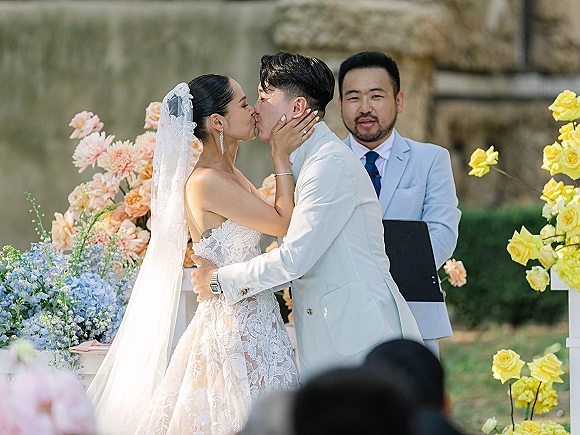 Wedding kiss as bride in lace dress and long veil holds groom’s face, his white suit boutonniere, in a garden ceremony by a stone wall