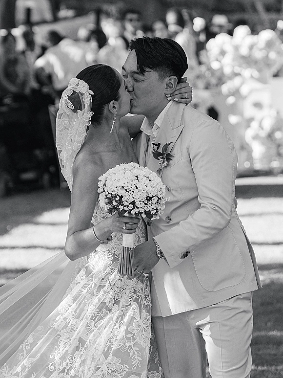 Wedding kiss portrait of bride and groom kissing, her lace dress and bouquet visible, with garden guests softly blurred behind them
