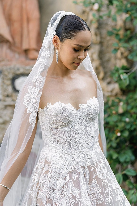Bridal portrait of a bride looking down in side profile, wearing a strapless lace wedding dress on outdoor steps by a stone wall and greenery