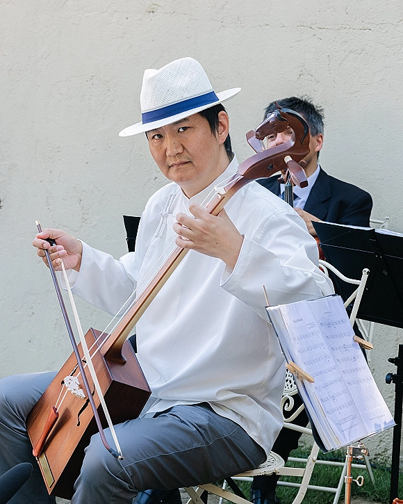 Wedding musician cellist playing with bow beside a music stand, wearing a white fedora and shirt on an outdoor lawn near others
