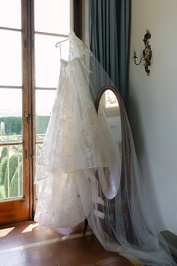 Wedding dress, lace wedding dress hanging on a hanger with a tulle veil draped beside an oval mirror near a tall window with blue curtains