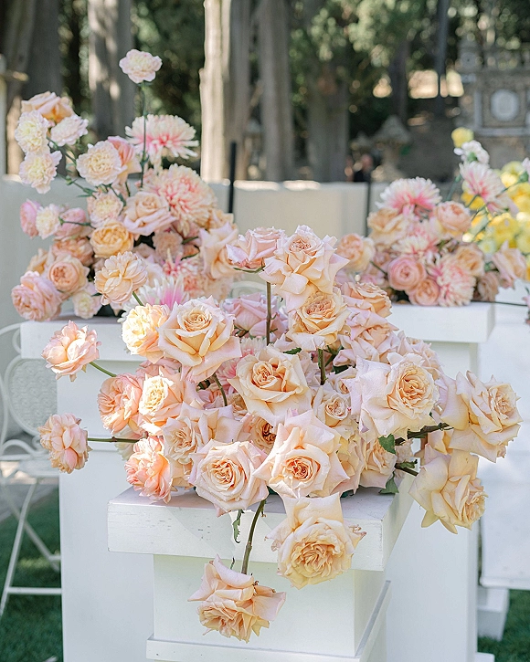 Wedding floral arrangement with peach rose wedding flowers and blush blooms on white pedestal stands set along a garden lawn with trees