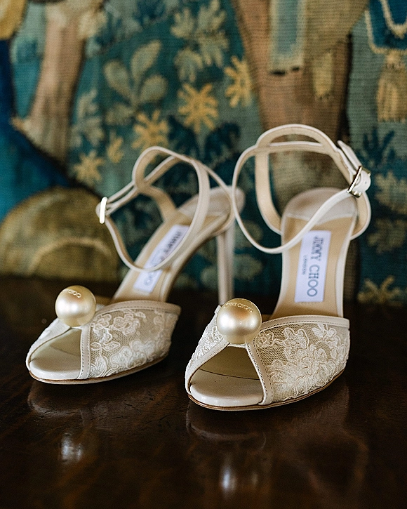 Bridal shoes with lace wedding heels styling, featuring pearl embellishments and ankle straps on a patterned chair above a wooden floor