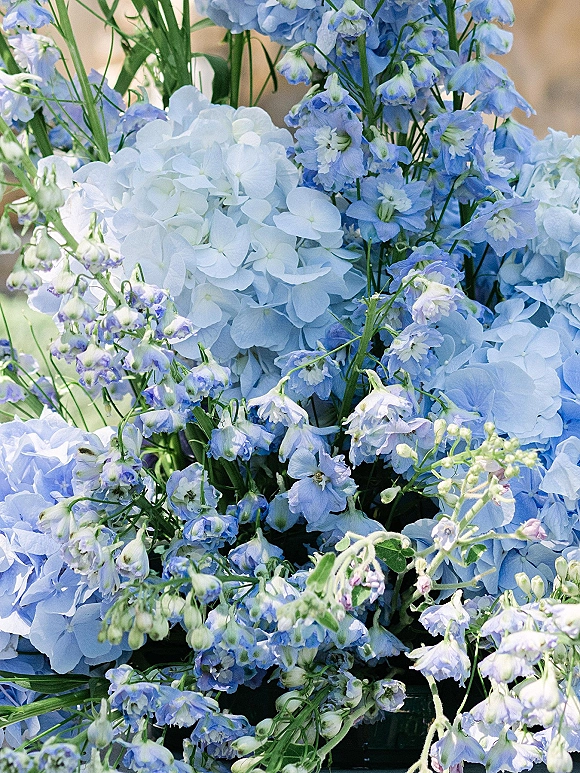Wedding flowers in a blue hydrangea centerpiece with delphinium and greenery accents on an outdoor lawn with a softly blurred garden backdrop