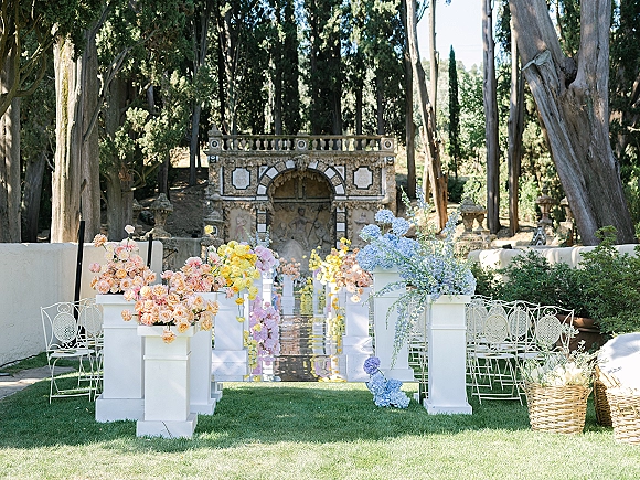 Ceremony aisle decor with a mirror aisle runner lined by white pedestals and pastel hydrangea, rose, and delphinium florals in a garden lawn setting