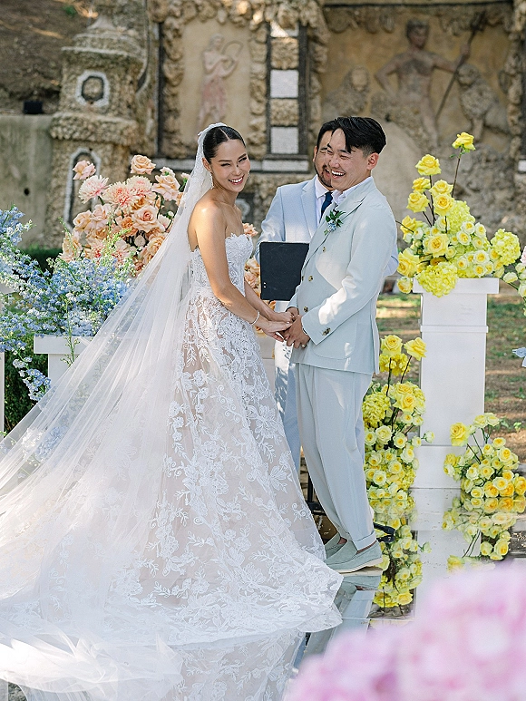 Ceremony moment as bride and groom hold hands laughing, her lace dress and veil beside his light blue suit on a mirrored aisle