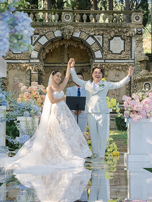 Ceremony moment as bride and groom raise hands in celebration, walking a reflective aisle beneath a stone arch with florals behind them