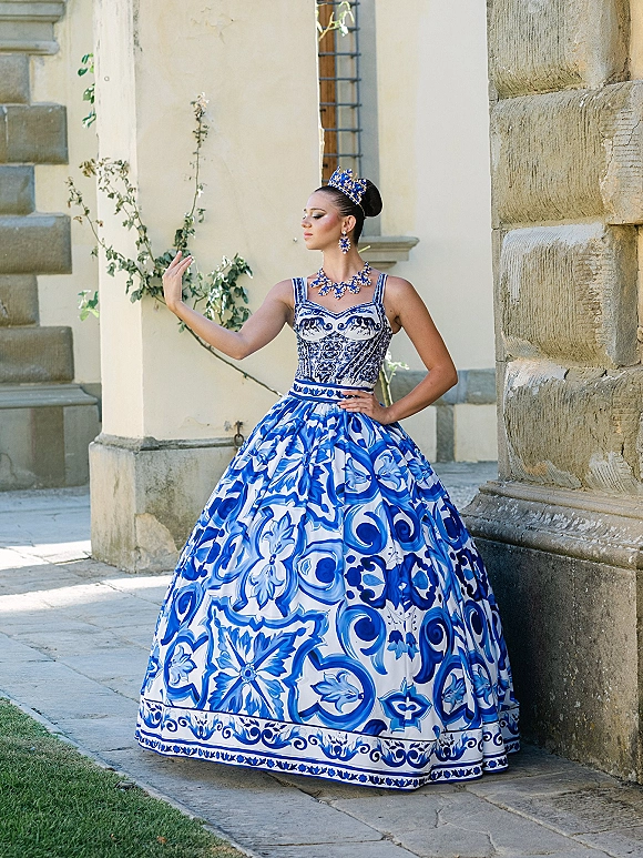 Bridal portrait in a blue and white wedding dress, wearing a tiara and statement necklace in a vine-lined courtyard with stone columns