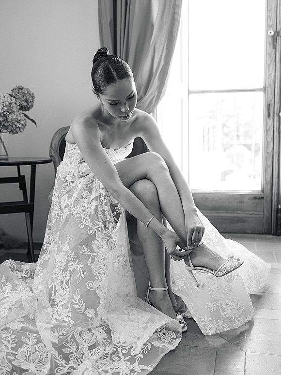 Bride getting ready, adjusting ankle strap heels on a chair in a strapless lace wedding dress, lit by window light and curtains
