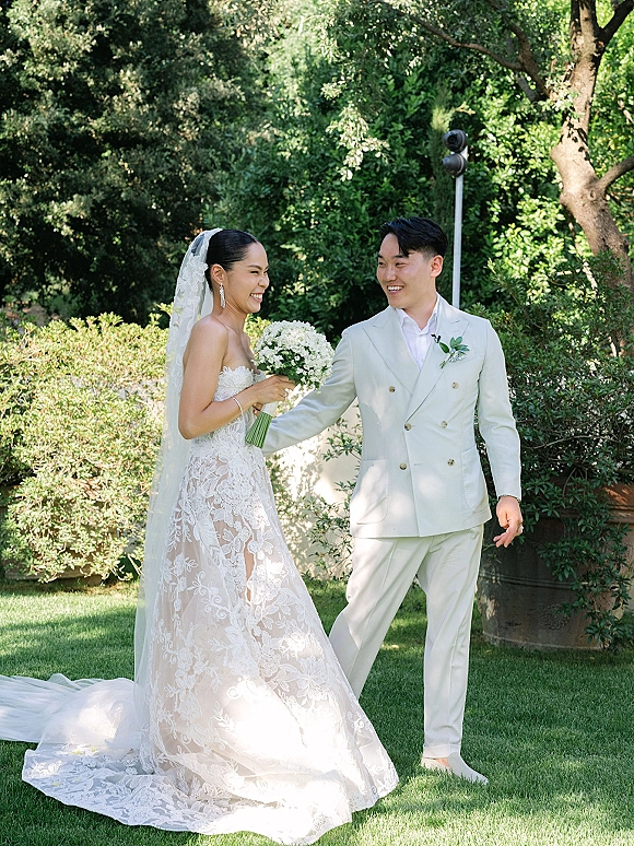 Couple portrait of bride holding a white bouquet in a strapless lace dress and long veil beside groom in white suit on a garden lawn