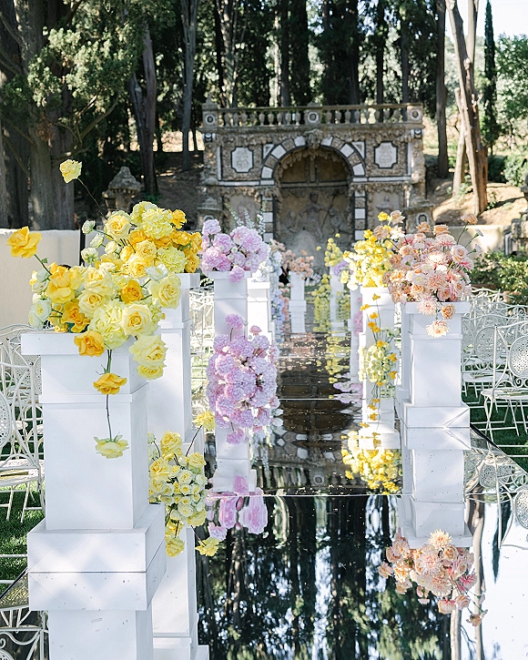 Ceremony aisle decor with a mirror aisle runner lined by white pedestal plinths and pastel florals on a garden lawn by stone fountain