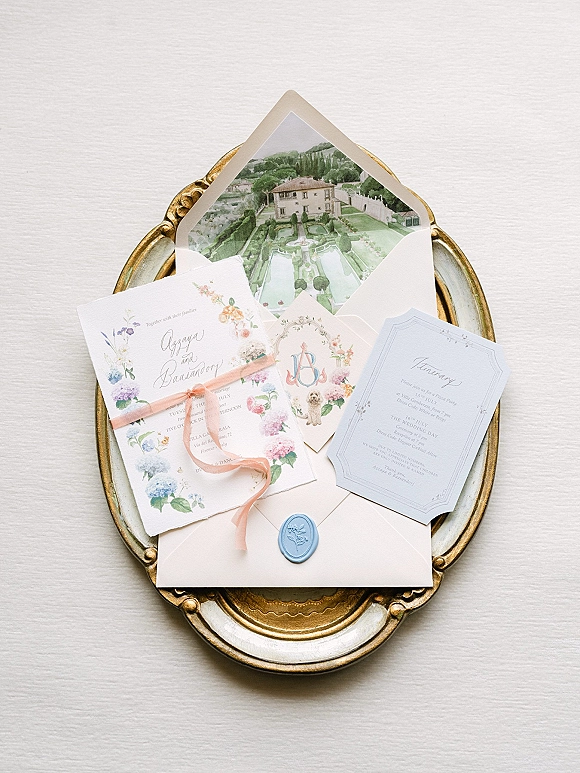 Invitation suite with floral wedding invitations, RSVP and itinerary cards, blush ribbon and wax seal arranged on a vintage gold tray over linen