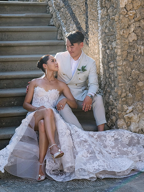 Couple portrait of bride and groom sitting on steps, groom’s arm around her as she looks up, lace dress train on stone staircase