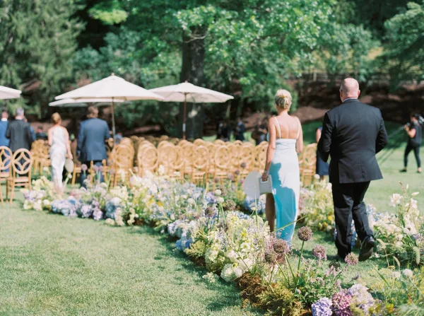 Wedding processional with bridesmaid walking down aisle past hydrangea arrangements on a grass lawn, as guests watch under white parasols